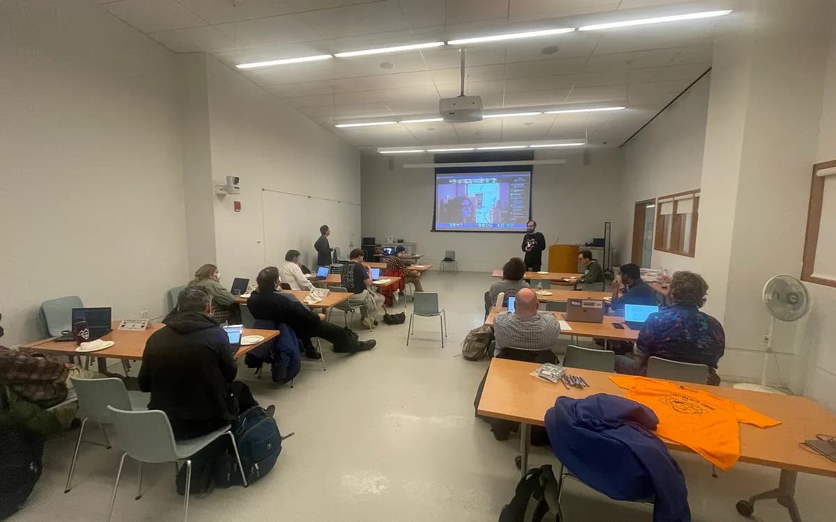 A hybrid meeting in a library room: people at tables with laptops while remote participants appear on a projected video call