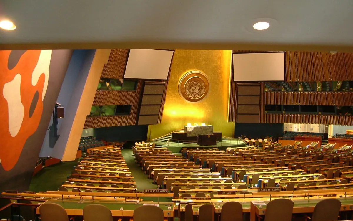 Rows of empty delegation desks on the floor of the UN General Assembly Hall, facing the gold wall and UN emblem behind the podium
